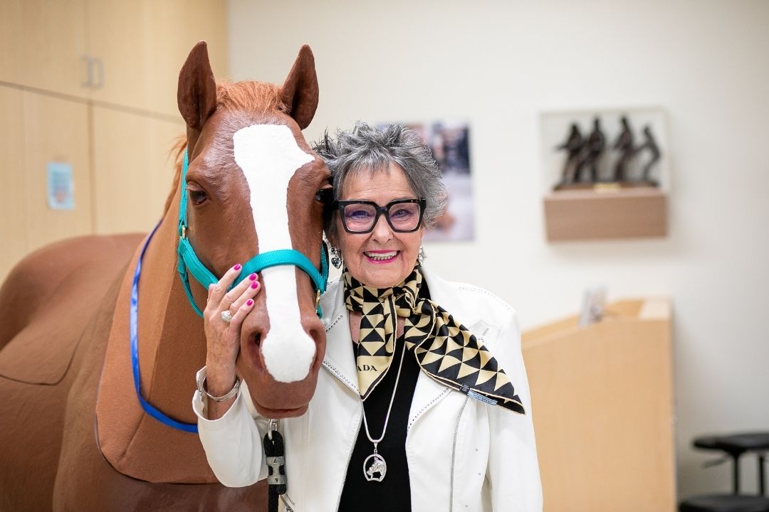 A woman with short hair and glasses stands next to a simulated horse.