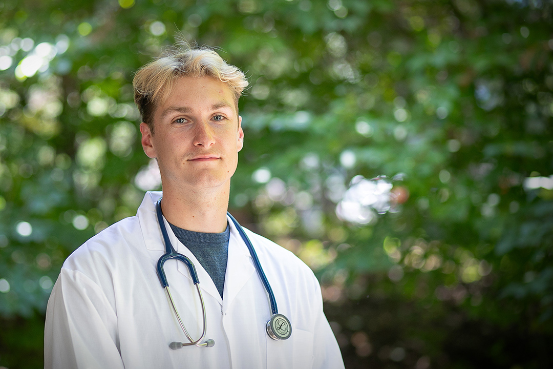 A veterinary student (Chad Steverding) in a white lab coat stands in front of green trees.